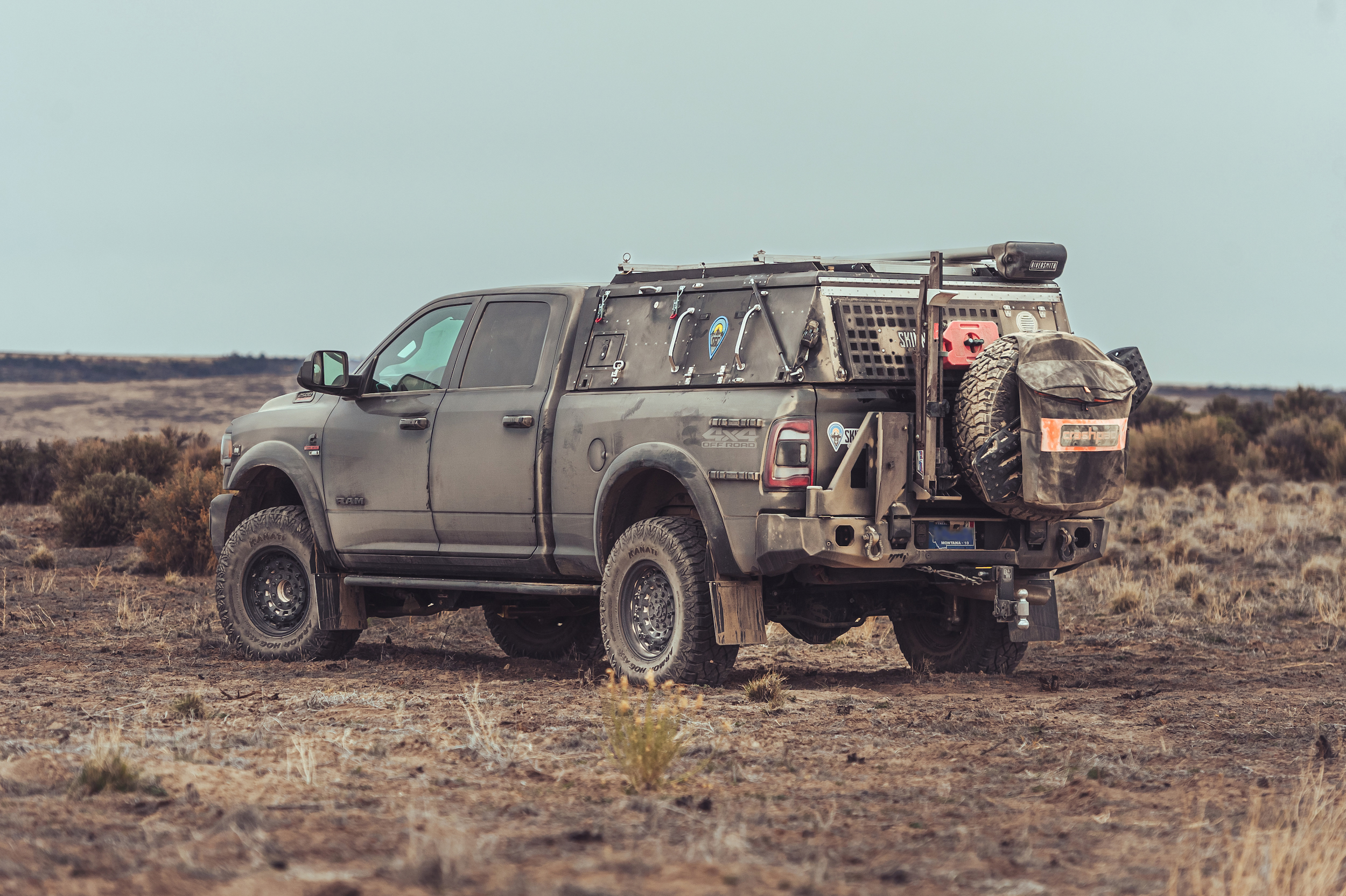 A rugged off-road truck parked on a barren, rugged landscape. This overland beast is equipped with large tires, a spare tire at the rear, jerry cans, and various gear attached to the bed. The terrain is a mix of dirt and sparse vegetation under an overcast sky.