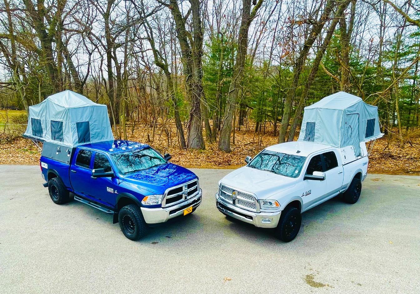 Two pickup trucks, outfitted for overland adventures, are parked next to each other in a forested area. Each truck has a rooftop tent set up. The truck on the left is blue, while the one on the right is white. The background features trees with minimal foliage and a ground covered in dry leaves.