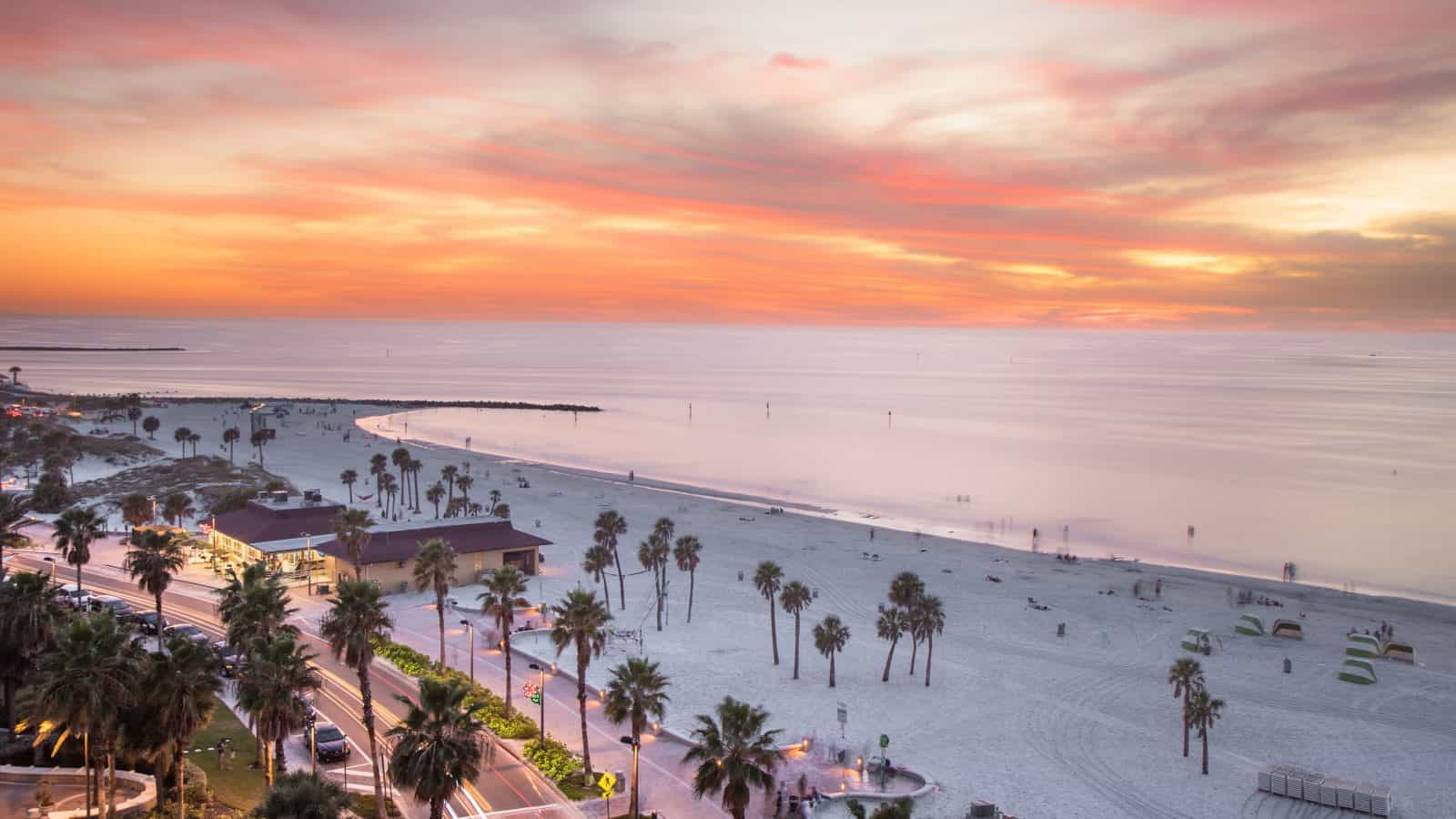 A scenic beach at sunset with a colorful sky displaying shades of orange, pink, and yellow. The sandy shoreline is dotted with palm trees and small shelters perfect for campers. A road runs parallel to the beach, bordered by more palm trees and lighted buildings. The calm ocean reflects the sunset.