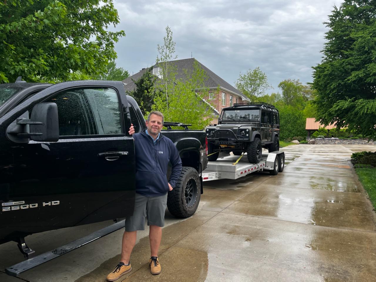 A man stands holding the driver's side door of a large black pickup truck parked on a wet driveway. The truck, ideal for overland adventures, is hitched to a trailer carrying a black off-road vehicle. Trees and a brick house are visible in the background. The sky is overcast.

