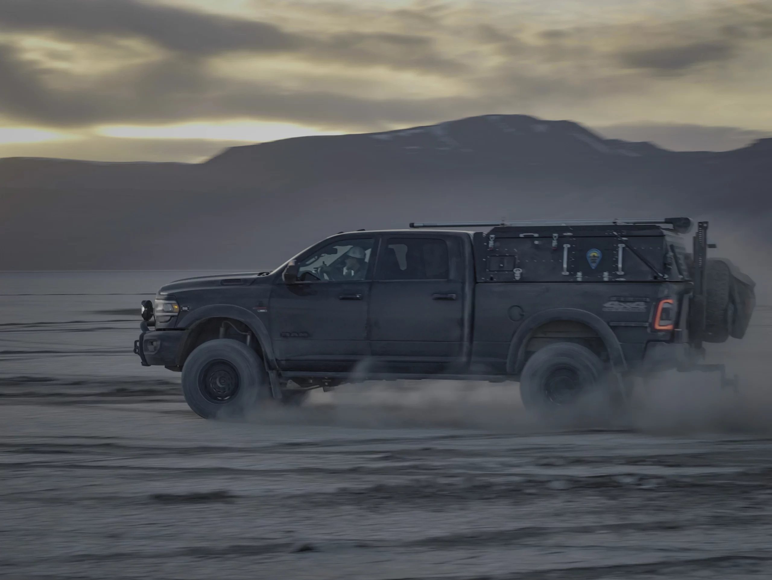 A rugged, black pickup truck drives across a barren, dusty landscape with mountains in the background at dusk. The vehicle, adorned with Skinny Guy branding and equipped for an overland expedition, appears ready for adventure.