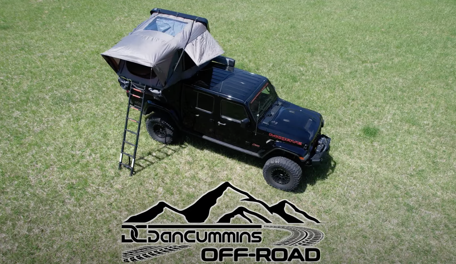 Aerial view of a black off-road vehicle with a rooftop tent set up on a grassy field. The tent is open, with a ladder leading up to it, perfect for overland adventurers. The Dan Cummins Off-Road logo, featuring mountains and a tire tread pattern, is displayed at the bottom of the image.