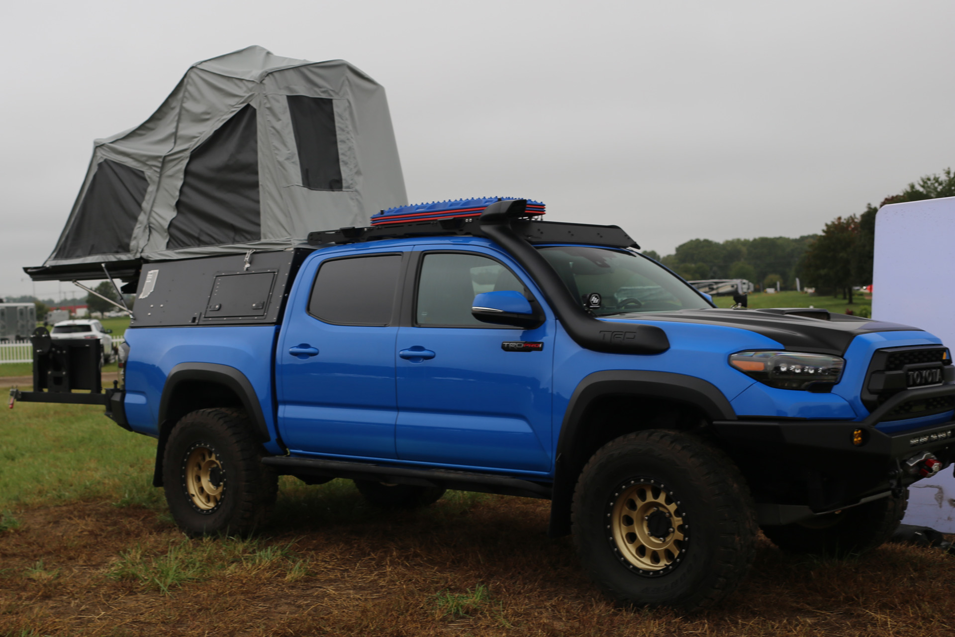 A blue Skinny Guy off-road truck is shown with a rooftop tent set up. The truck has rugged tires, a roof rack with additional storage, and various modifications for overlanding. The background features a grassy area with some trees and other campers.